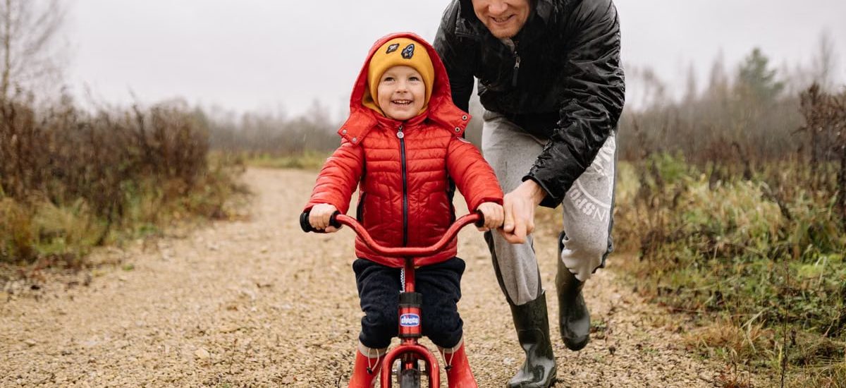 A father joyfully helps his child ride a bicycle on a gravel path in an autumn setting.