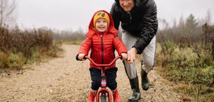 A father joyfully helps his child ride a bicycle on a gravel path in an autumn setting.