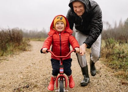 A father joyfully helps his child ride a bicycle on a gravel path in an autumn setting.